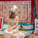 Woman in traditional attire sits amidst ornate textiles.