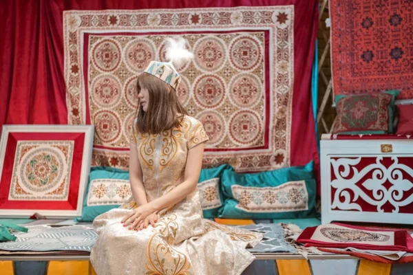 Woman in traditional attire sits amidst ornate textiles.